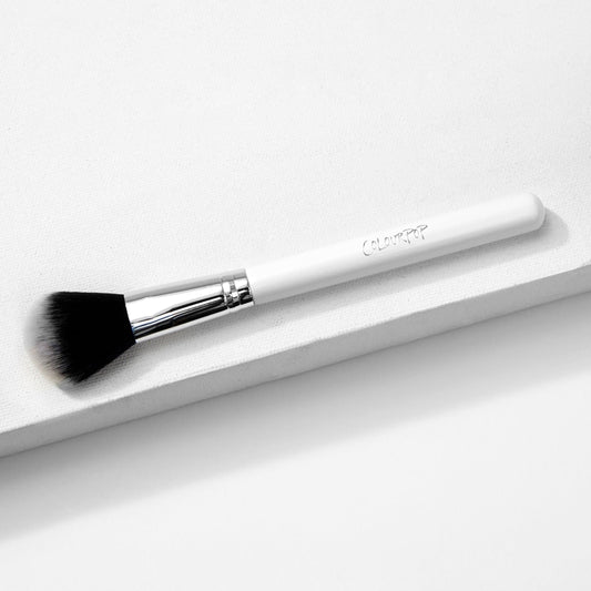 Blush Brush - ColourPop Makeup Brush with soft black-to-white bristles and chrome ferrule - lying diagonally on a white textured platform, white handle stamped ColourPop, ideal for sweeping and blending powder blush.
