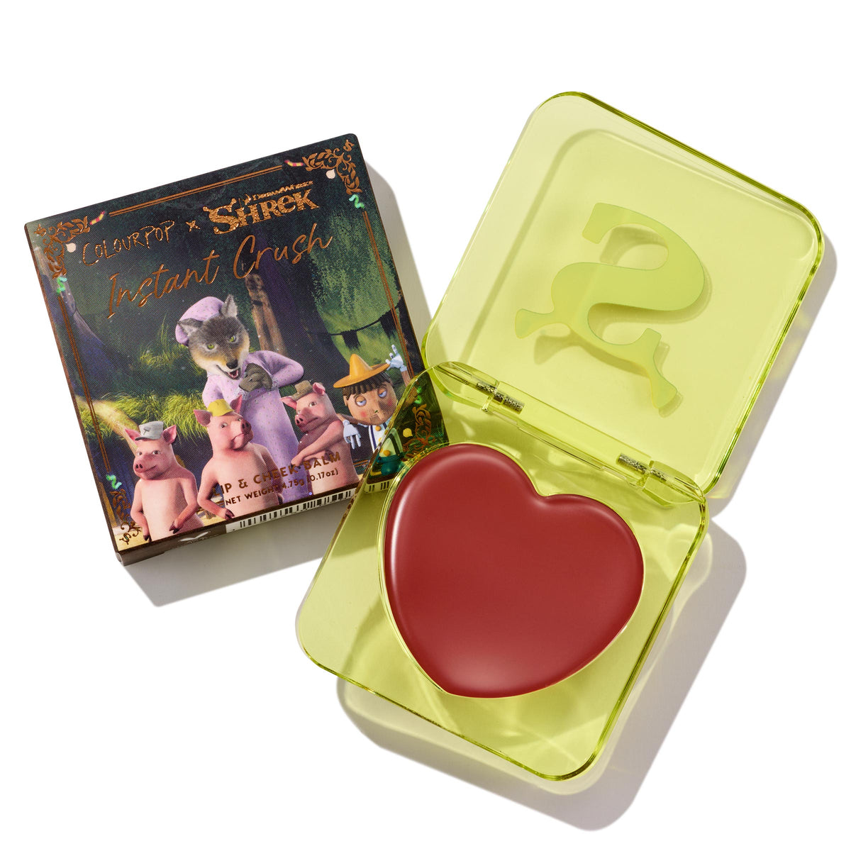Fairytale Things ColourPop Lip and Cheek Balm sits open in a translucent neon-yellow square compact with an embossed S, revealing a red heart-shaped pan; beside it, the Shrek collab carton shows fairytale characters on a white studio background.