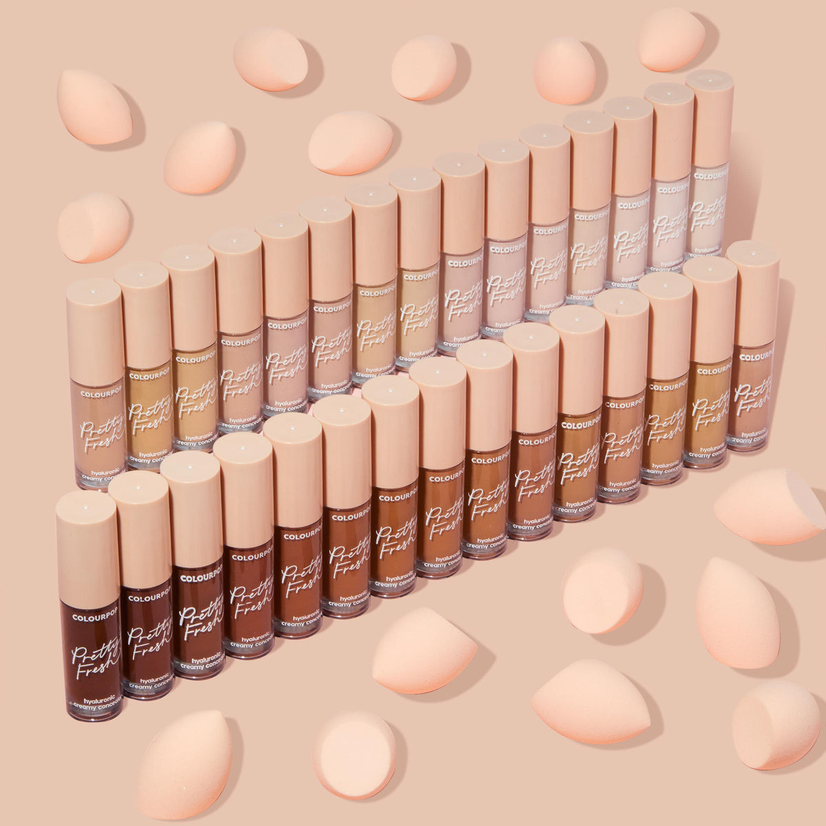 ColourPop Pretty Fresh Creamy Concealer, shade Light 50 W, standing with the full shade range in neat rows; frosted tubes with blush-pink caps and peach blending sponges scattered on a nude background.