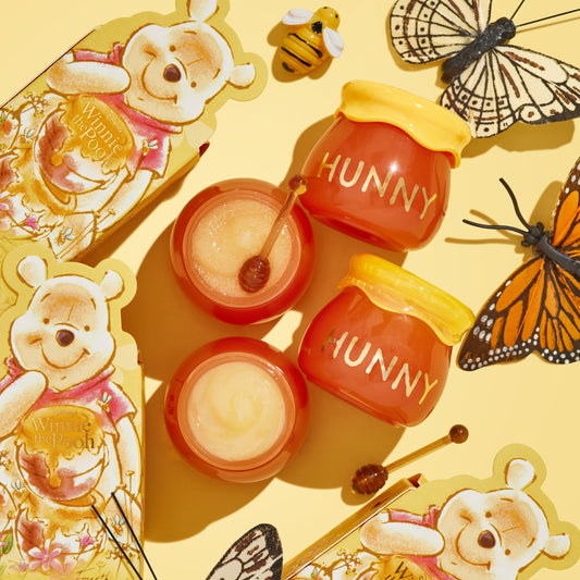 Sweet as Hunny ColourPop Eye & Lip Set displayed in a flat lay, featuring honey pot-shaped jars labeled HUNNY, two open pots of shimmery balm with mini honey-dipper applicators, and Winnie the Pooh packaging, butterflies, and a bee on a yellow background.