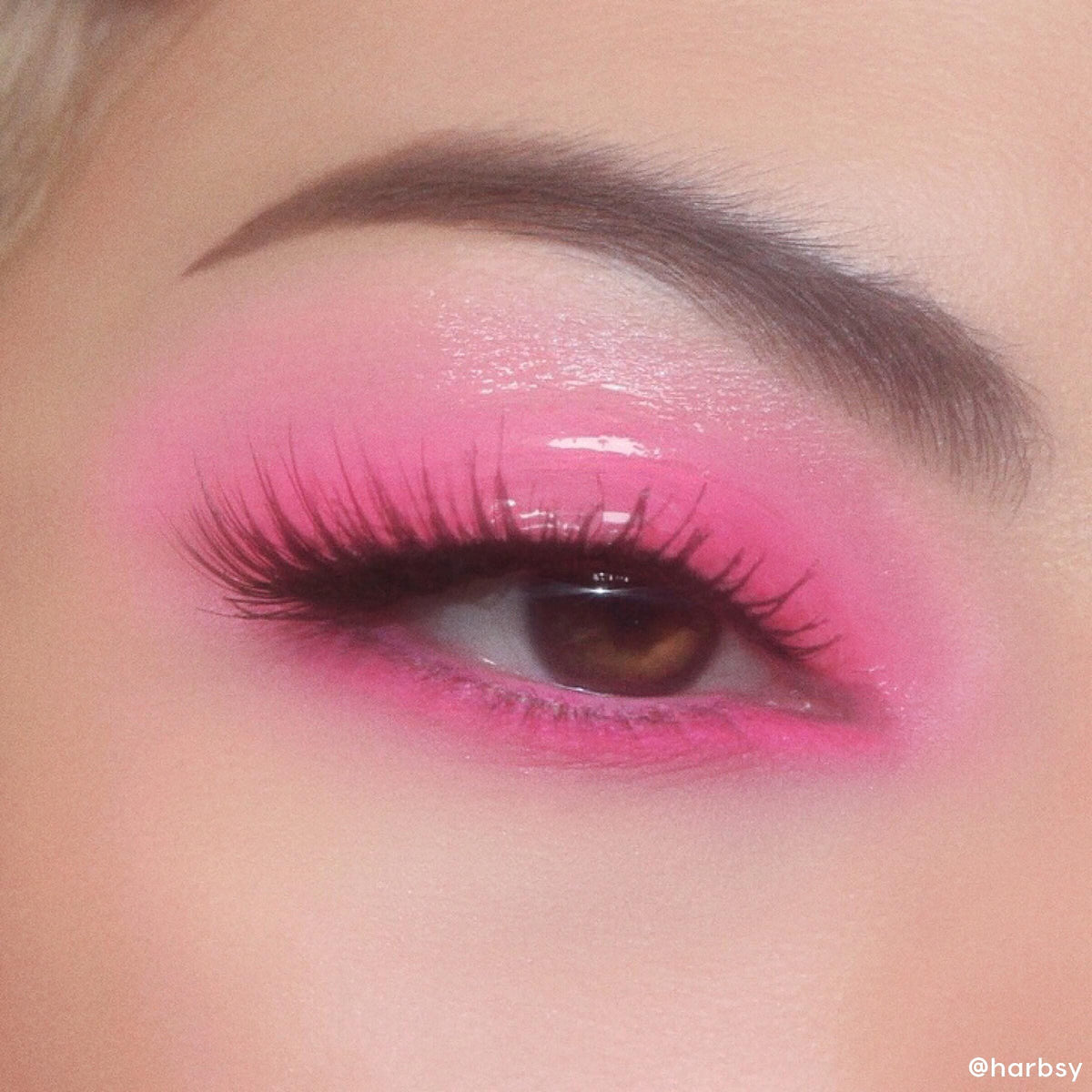 Glossy eyelid wearing In The Clear Face & Eye Gloss layered over vibrant pink eyeshadow, creating a glassy wet-look on a brown eye with long lashes and a defined brow; ColourPop Face & Eye Gloss product detail.