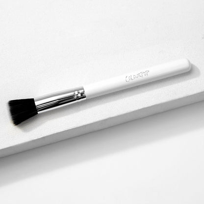 ColourPop stippling makeup brush resting diagonally on a white textured block, featuring duo-fiber black-and-white bristles, a shiny silver ferrule, and a white handle with the ColourPop logo.