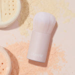 Mini Kabuki Face Brush buffing loose setting powder, a short-handled ColourPop Makeup Brush with dense white bristles and a blush-pink barrel, resting amid peach and yellow powders beside open sifter jars, ideal for travel and seamless blending.