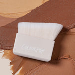 ColourPop Makeup Brush Angled Sculpting Brush resting on swatches of tan to deep brown cream contour; dense white angled bristles extend from a beige block handle stamped ColourPop, ideal for precise cheekbone sculpting.