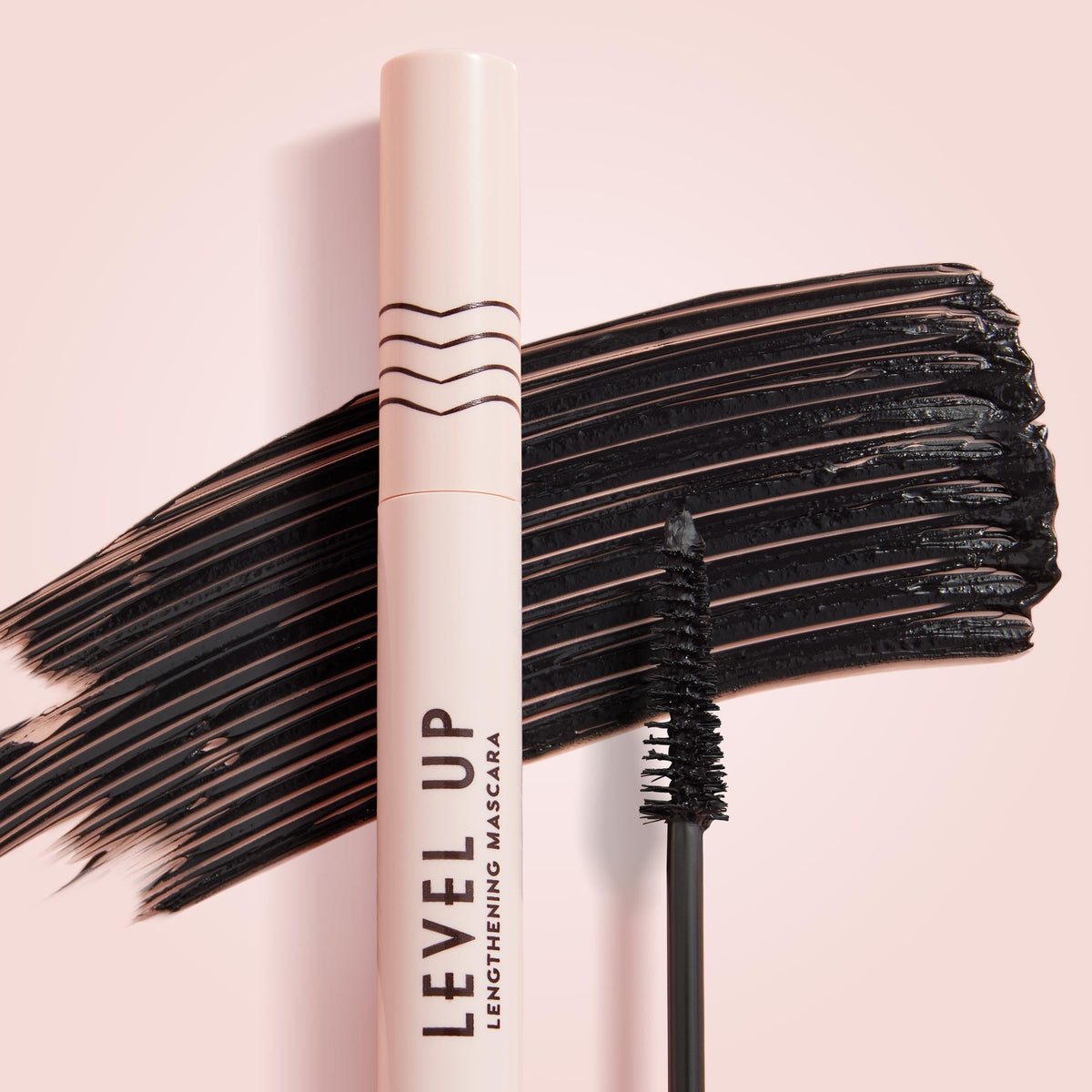 ColourPop Lengthening Mascara Level Up in Black stands upright with its wand coated in pigment, set before bold black swipes on a soft pink background, highlighting the slim brush and blush-pink tube for dramatic lash length.