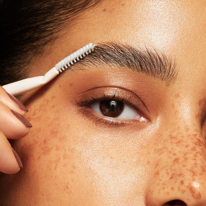 Brow brush sweeping eyebrow hairs upward for a feathered finish, close-up of an eye with freckled skin; white angled spoolie makeup brush with ergonomic handle from ColourPop, shaping a clean, natural brow arch.