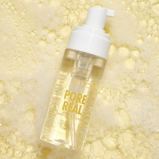 Pore Real Cleanser foaming pump bottle with white cap and clear liquid, gold “Pore Real” label, resting on glistening yellow lather and bubbles, highlighting ColourPop Face Cleanser by Fourth Ray Beauty for a pore-cleansing wash.