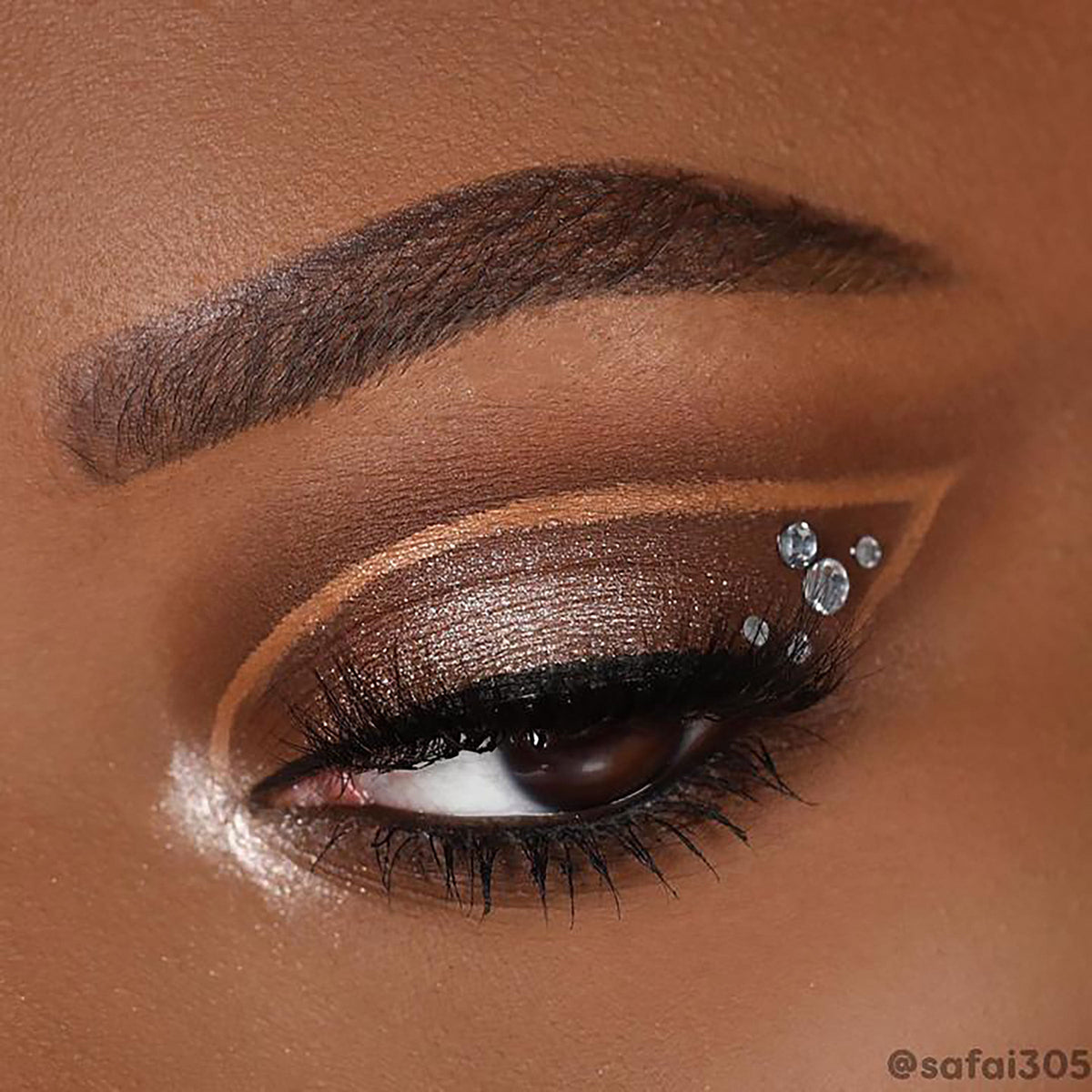 Shimmery champagne-bronze cut-crease eye makeup created with the ColourPop Feelin' Bubbly shadow palette, winged shape with black liner, long lashes, and three crystal studs at the outer corner in a tight close-up on the eyelid.