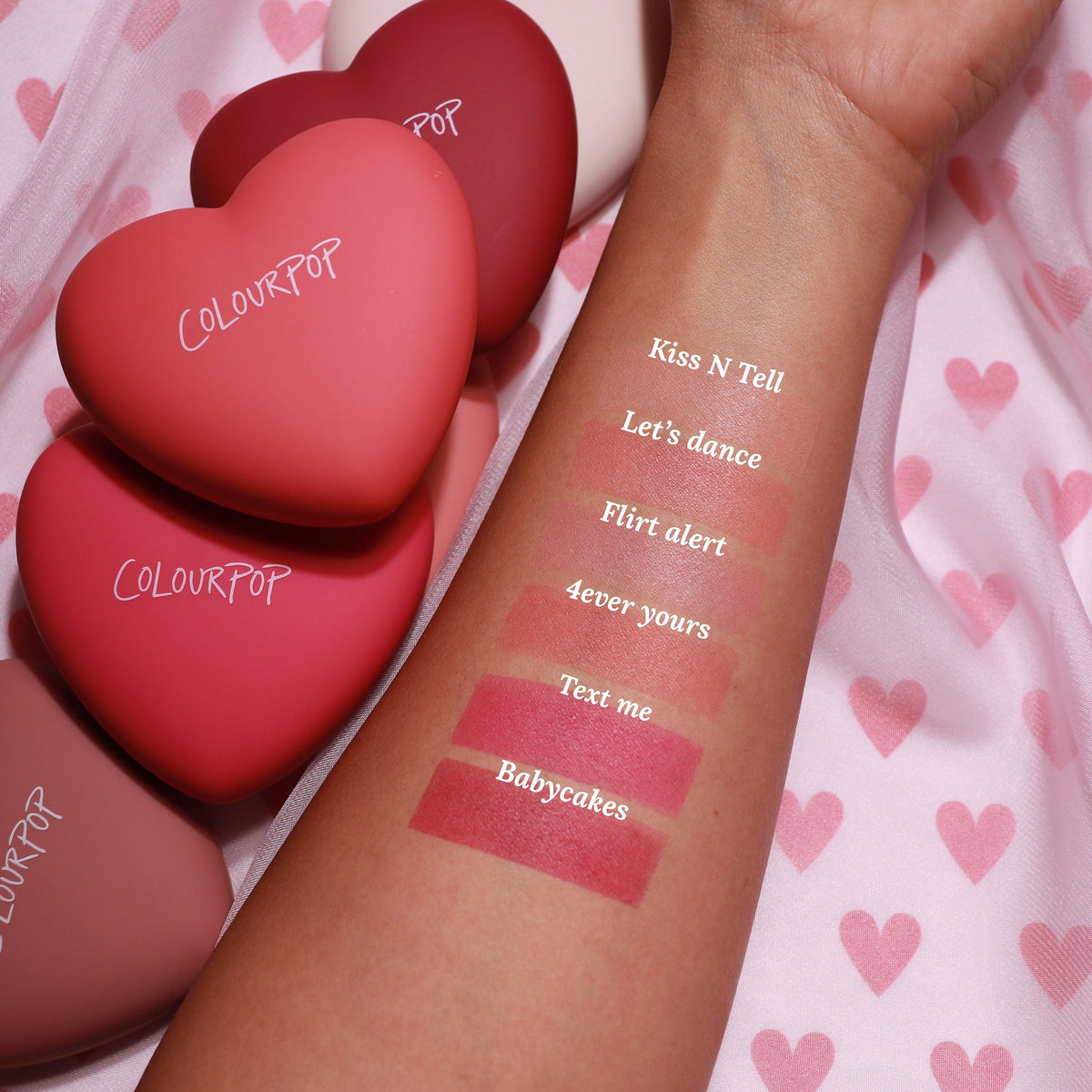 ColourPop Pressed Powder Blush Text Me swatched on a forearm, showing a pigmented rosy pink matte next to Babycakes, 4ever Yours, Flirt Alert, Let’s Dance, and Kiss N Tell, with heart-shaped ColourPop compacts on pink heart-print fabric.