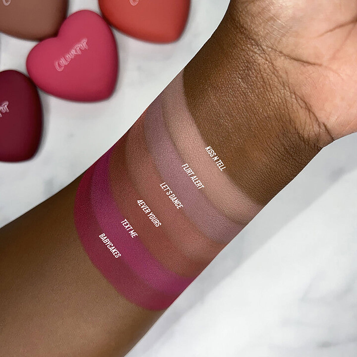 Babycakes swatch from ColourPop Pressed Powder Blush shown on deep skin, with labeled shade stripes - Babycakes, Text Me, 4Ever Yours, Let’s Dance, Flirt Alert, Kiss N Tell - showing rich pigment; heart-shaped ColourPop compacts blurred in the background.