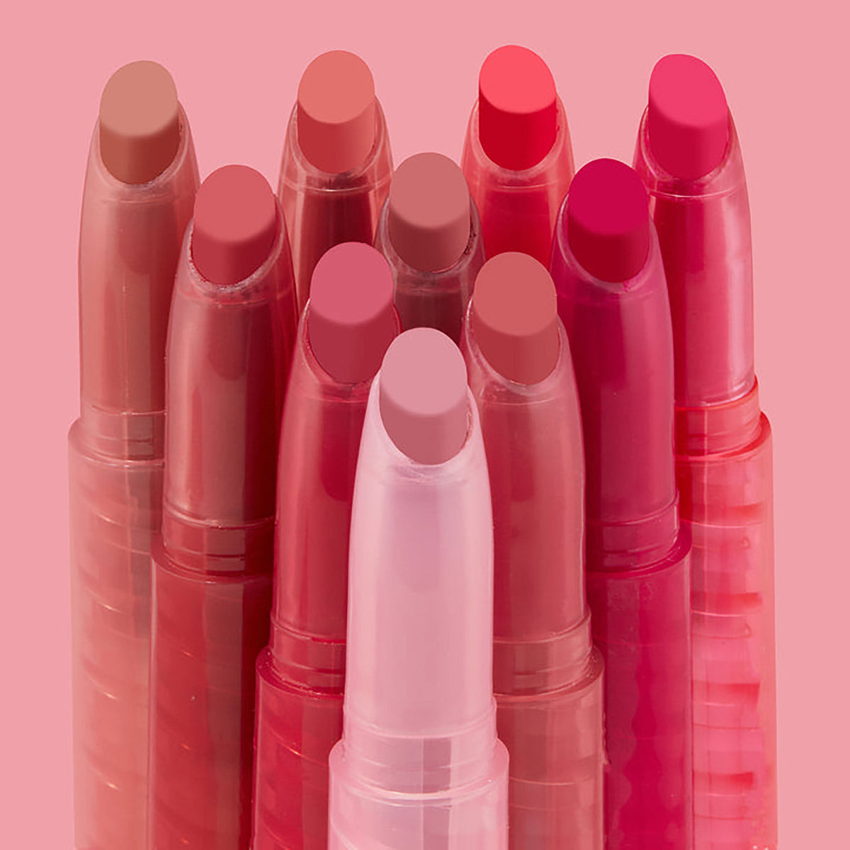 Just A Kiss ColourPop Lippie Stix set standing upright in a tight cluster, showcasing creamy bullet tips in nude, pink, coral, and berry tones, in translucent pink twist-up tubes against a soft blush background.