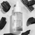 Glass dropper bottle of Fourth Ray Beauty All Clear Purifying Serum standing upright, showcasing clear formula and label, surrounded by charcoal pieces on a white marble surface; .85 fl oz/25 mL ColourPop Face Serum for purifying skin.