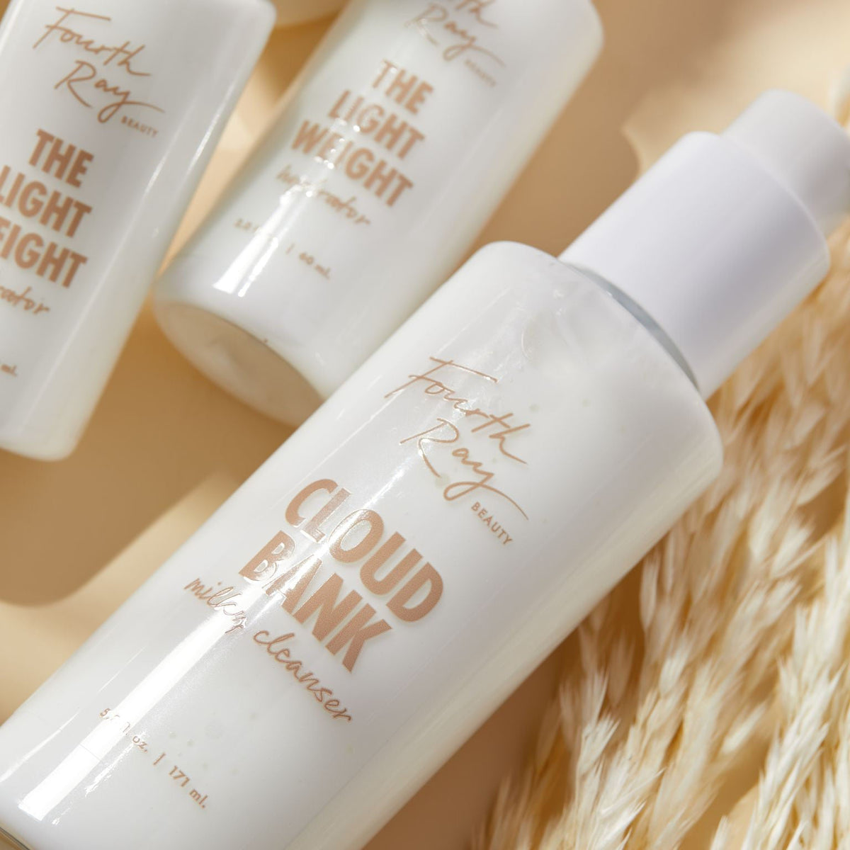 Fourth Ray Beauty Cloud Bank Milky Cleanser bottle resting diagonally with a white pump, creamy formula catching light; set on a warm beige surface beside dried grass with “The Light Weight” bottles blurred behind; ColourPop Face Cleanser, 5 fl oz.