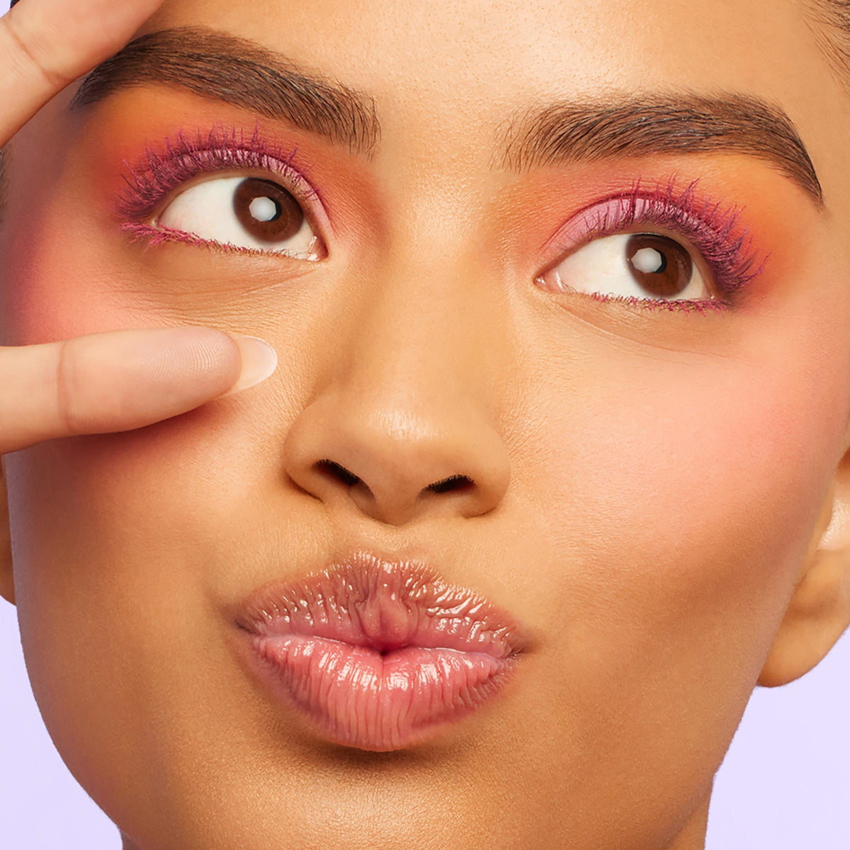 Lashes coated in vivid hot-pink ColourPop BFF Mascara, shade Pink Inc., fan out on both eyes as a model puckers glossy lips and frames one eye with fingers, a soft lavender backdrop highlighting the bold length and color payoff.