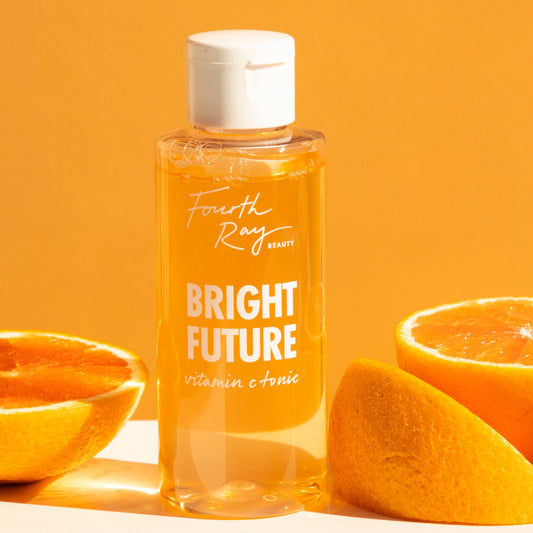 Bright Future Vitamin C Tonic bottle by Fourth Ray Beauty standing upright, showing golden liquid with bubbles and a white flip cap, flanked by fresh orange halves on a sunny orange backdrop; ColourPop vitamin C skincare brightening tonic.