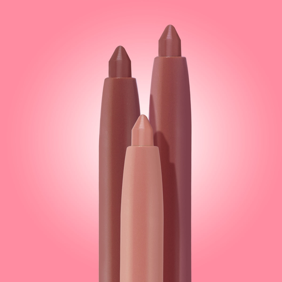 Three chubby lip liners in nude, rose, and mauve stand upright with pointed tips against a pink gradient backdrop, highlighting the Lips Don’t Lie ColourPop So Juicy Plumping Lip Liner Set, a trio for precise, plumped-looking lips.