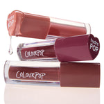 Three ColourPop Lip Stain tubes from the Everyday Besties set stacked on a glossy white surface; the top nude shade drips a clear droplet while mauve and rose tones sit below, clear barrels and a heart-shaped hydraPOP cap highlight the stain formula.