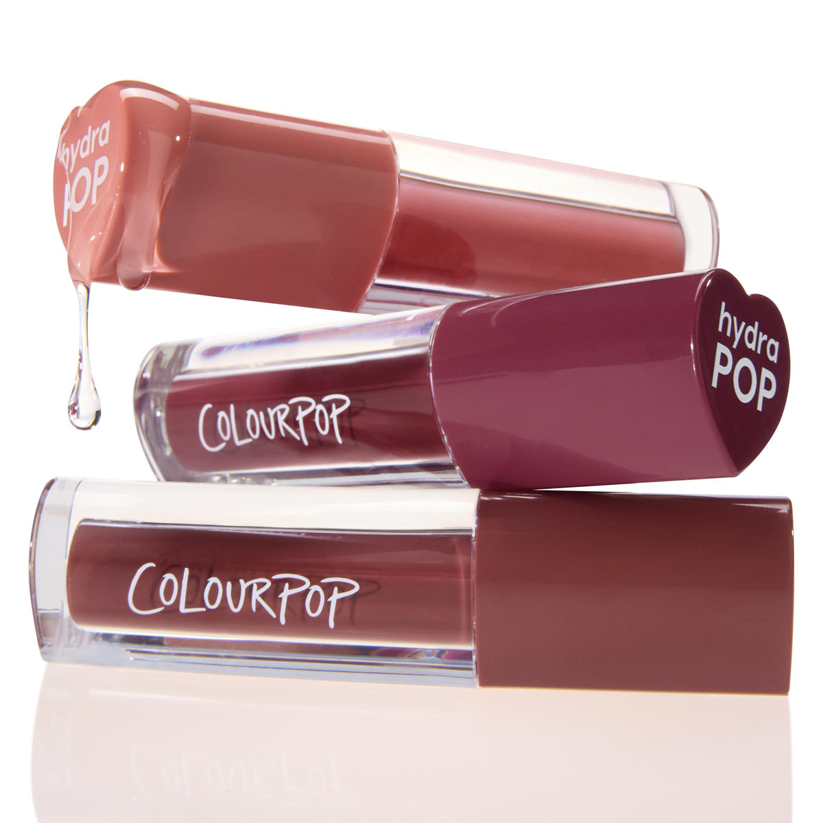 Three ColourPop Lip Stain tubes from the Everyday Besties set stacked on a glossy white surface; the top nude shade drips a clear droplet while mauve and rose tones sit below, clear barrels and a heart-shaped hydraPOP cap highlight the stain formula.