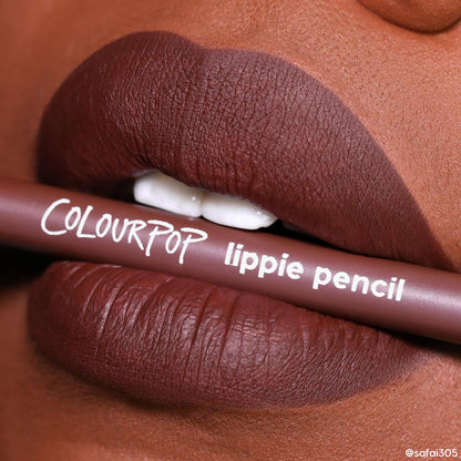 ColourPop Lippie Pencil, shade Pitch, held between full matte deep-brown lips, showcasing a crisp defined lip line and rich chocolate color in a close-up beauty shot.