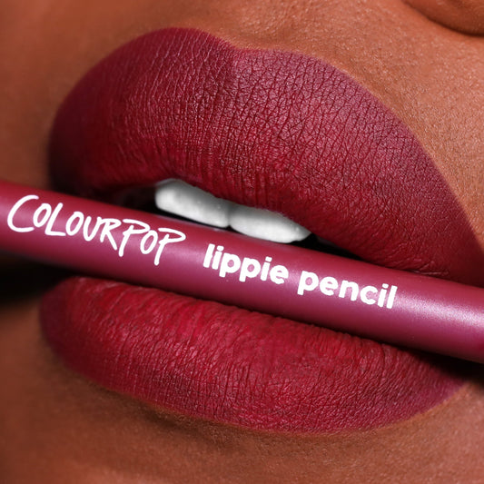 ColourPop Lippie Pencil in On Ice held between matte, berry-red lips, showcasing precise lip definition; mauve pencil with white logo in a tight beauty close-up, highlighting the liner's rich pigment and velvety matte finish.
