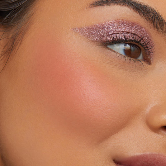 A-Lister ColourPop Cream Blush delivering a soft peachy-coral flush on the cheekbone with a dewy finish, shown in a close-up of a model’s face paired with shimmering mauve winged eyeshadow and defined lashes in natural light.