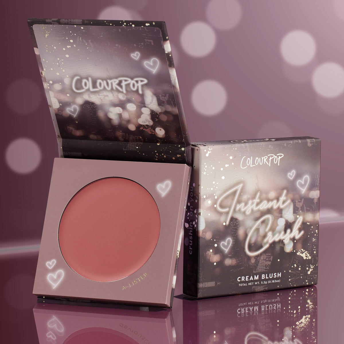 A-Lister cream blush compact sits open to reveal a warm rosy-mauve pan, with the matching box beside it; ColourPop Cream Blush Instant Crush packaging features heart doodles and sparkles against a mauve bokeh backdrop on a glossy surface.