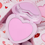 Cupid’s Bow heart-shaped compact opens to reveal a cool baby-pink pan and mirror, showcasing the ColourPop Pressed Powder Blush amid Valentine-themed boxes with hearts and lips, highlighting a soft, romantic cheek color.