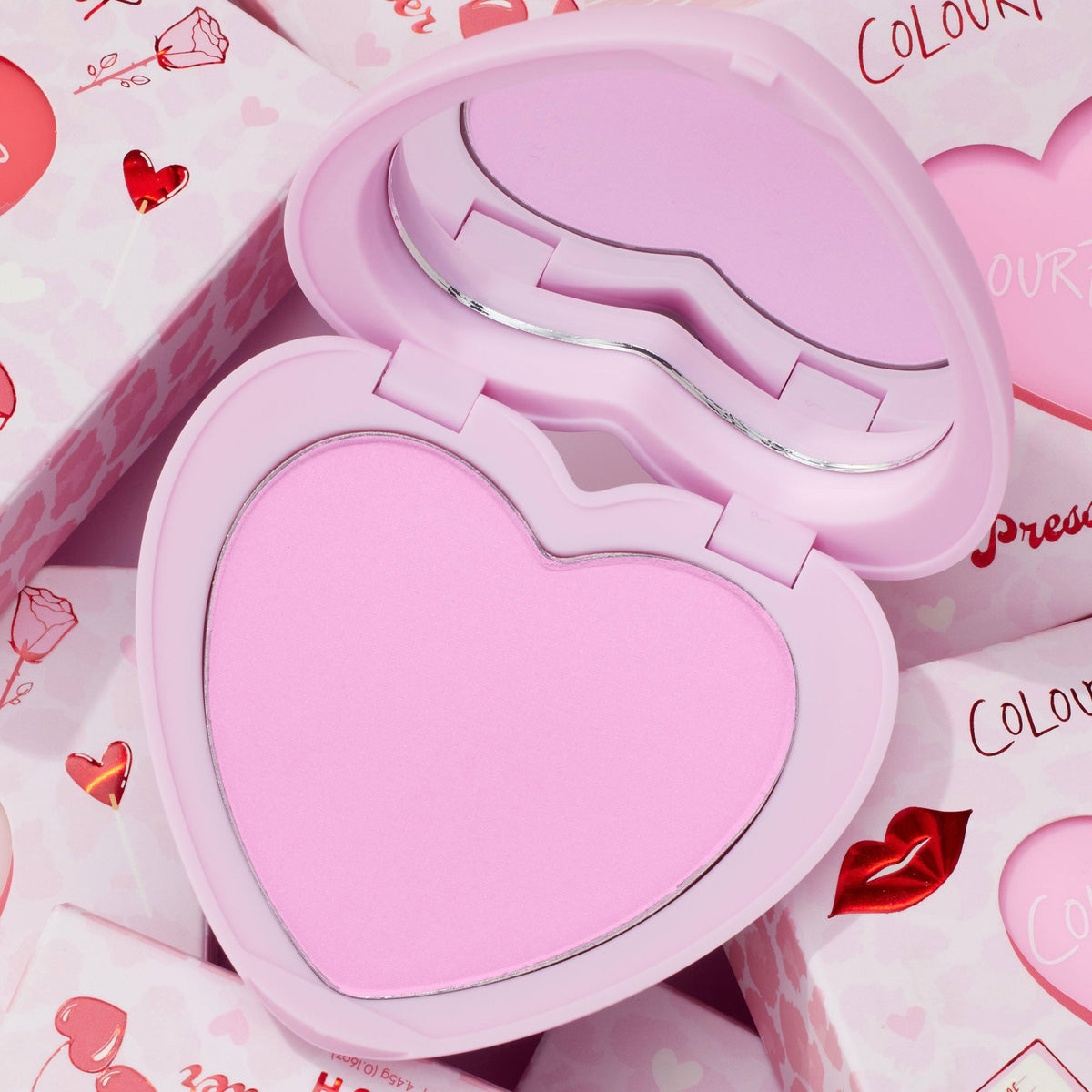 Cupid’s Bow heart-shaped compact opens to reveal a cool baby-pink pan and mirror, showcasing the ColourPop Pressed Powder Blush amid Valentine-themed boxes with hearts and lips, highlighting a soft, romantic cheek color.