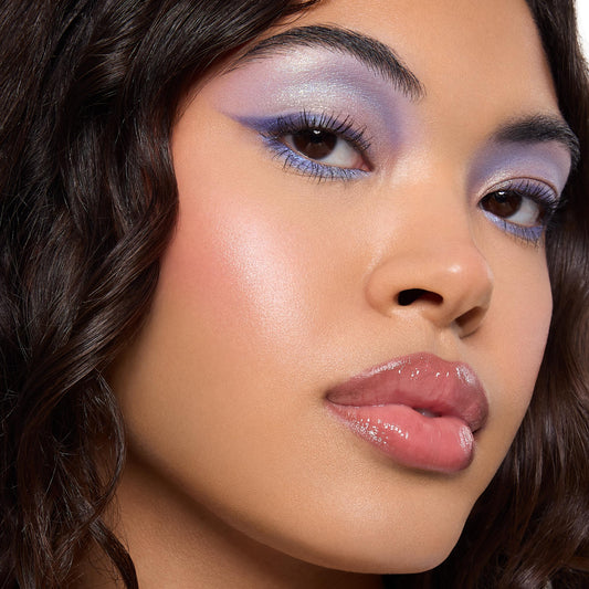 Model shows icy lavender and periwinkle winged eyeshadow with blue lower‑lash liner and silver shimmer, showcasing the Beach Dreamin' ColourPop Shadow Palette; dewy glow, pink blush, and glossy nude lips in a close‑up studio beauty shot.