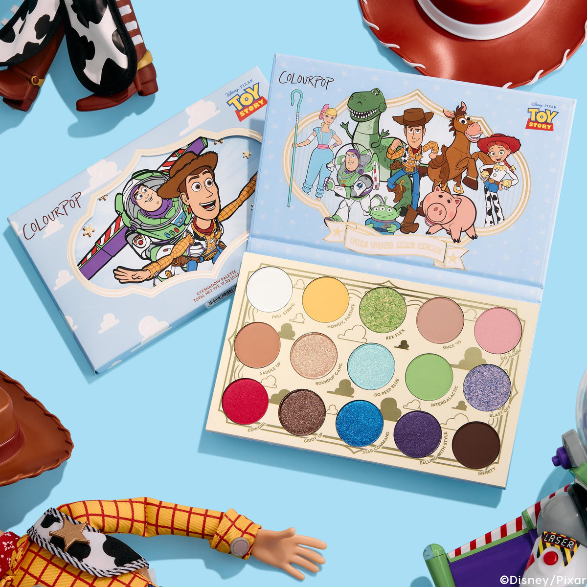 ColourPop The Toys Are Here shadow palette lays open, displaying 15 matte and shimmer shades - white, yellow, greens, blues, purple, pink, red, and neutrals - beside Toy Story character cover art, styled top-down with dolls and a cowboy hat on a sky-blue backdrop.
