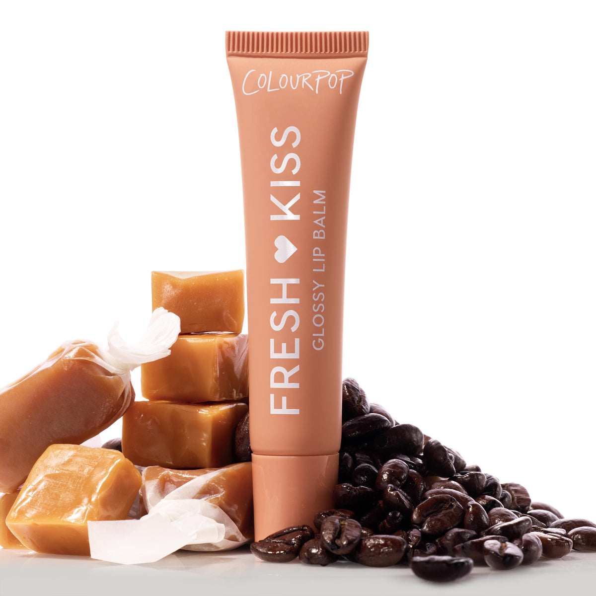 ColourPop Fresh Kiss Glossy Balm in Caramel Latte stands upright, a warm caramel tube with white “Fresh Kiss Glossy Lip Balm” lettering, surrounded by stacked caramel squares and glossy roasted coffee beans on a white backdrop.