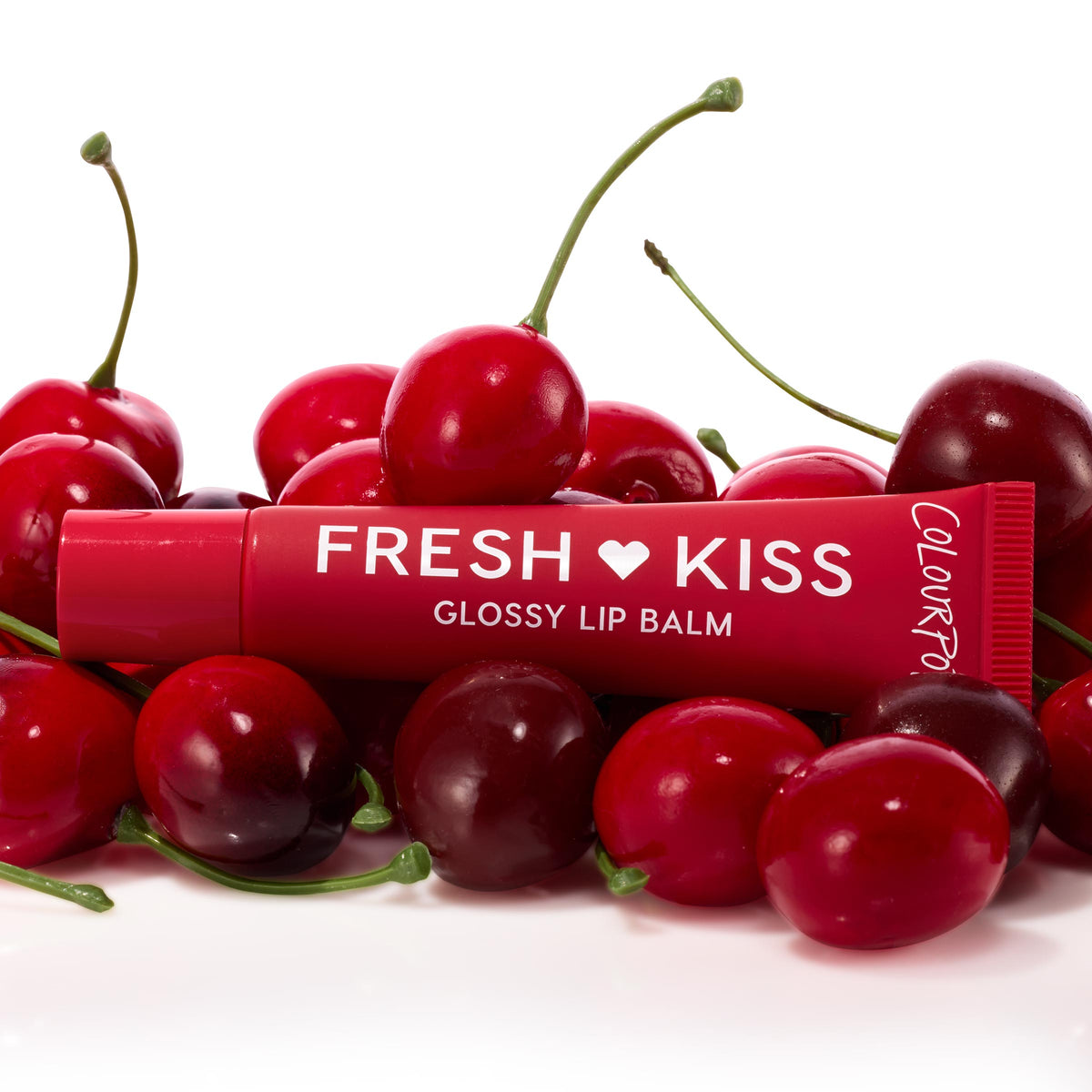 ColourPop Fresh Kiss glossy lip balm tube from the Get Rich ColourPop Glossy Lip Balm Trio resting among ripe cherries, showcasing a cherry-red tint and shiny finish in a studio close-up on a white background.