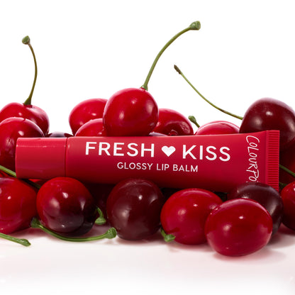 ColourPop Fresh Kiss glossy lip balm tube from the Get Rich ColourPop Glossy Lip Balm Trio resting among ripe cherries, showcasing a cherry-red tint and shiny finish in a studio close-up on a white background.