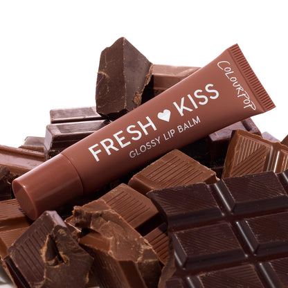 Fresh Kiss Glossy Lip Balm tube by ColourPop rests angled atop broken milk and dark chocolate bars, highlighting chocolate-brown packaging and white lettering; Sweet Treats ColourPop Glossy Lip Balm Trio product shot on a white studio backdrop.