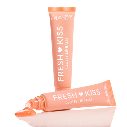 ColourPop Fresh Kiss Glossy Balm in Mango Sorbet, peach squeeze tube with white lettering, stands upright beside a second tube releasing a glossy peach gel onto a reflective white surface, highlighting the slanted applicator tip.