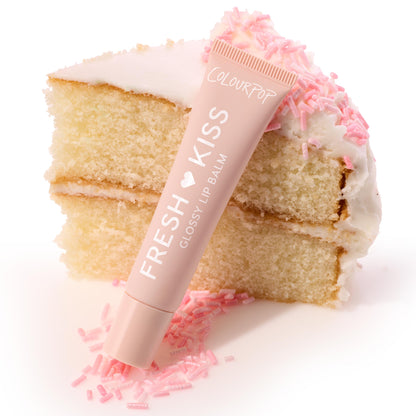 Pastel-pink ColourPop Fresh Kiss lip balm tube leaning against a slice of vanilla cake with white frosting and pink sprinkles, showcasing Pretty in Pastels, a ColourPop Glossy Lip Balm Trio.