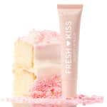 ColourPop Fresh Kiss Glossy Balm, Vanilla Cake, stands upright against a frosted vanilla cake slice topped with pink sprinkles, blush tube with white lettering styled on a bright, reflective surface for a sweet, high-shine lip finish.