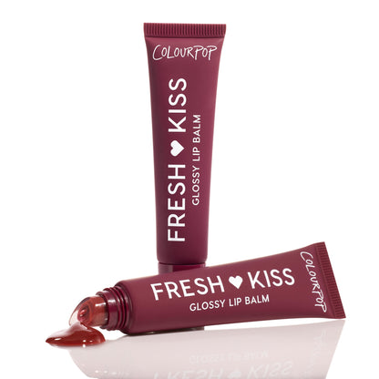 ColourPop Fresh Kiss Glossy Balm in Wild Berry stands upright while a second maroon squeeze tube lies uncapped with glossy berry tint pooling at the tip, on a white reflective studio background.
