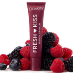ColourPop Fresh Kiss Glossy Balm in Wild Berry standing upright, deep berry tube with white lettering nestled among fresh raspberries, blackberries, and blueberries on a white backdrop, highlighting juicy, glossy lip care.