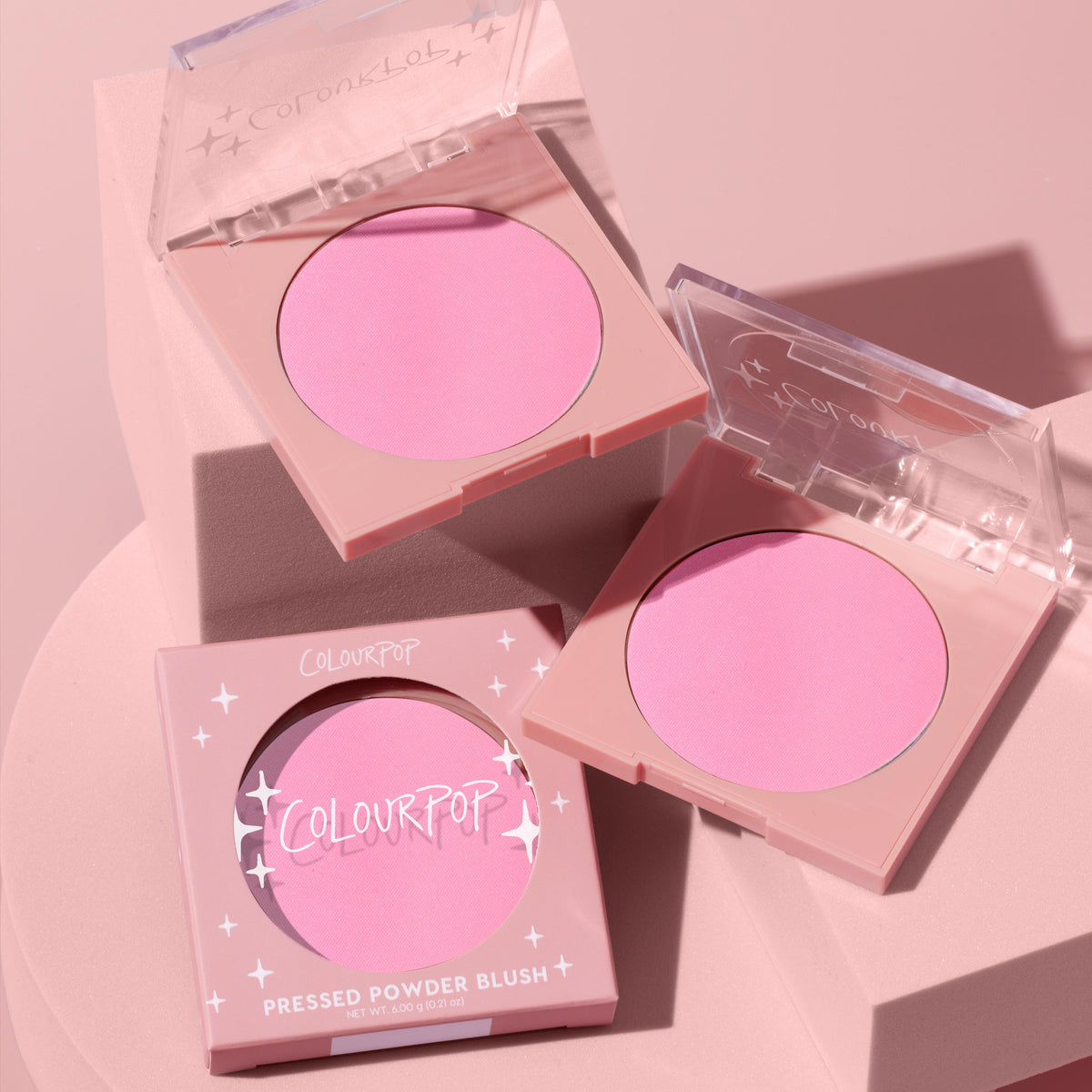 Flamingo ColourPop Pressed Powder Blush showing a vibrant bubblegum pink pan in two open square compacts with clear lids, plus one boxed unit, styled on a soft pink pedestal under studio lighting.