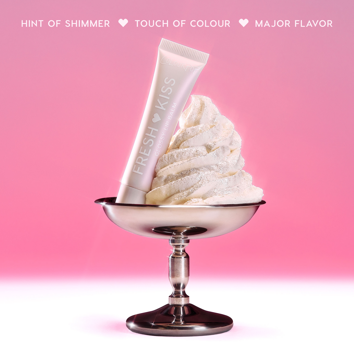 ColourPop Fresh Kiss Glossy Balm in Whipped Cream shade resting in a swirl of shimmering whipped cream in a silver dessert cup against a red gradient background, highlighting the glossy balm as a hybrid Lip Gloss & Lip Oil product.