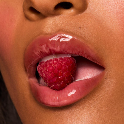 Glossy berry-tinted lips wearing ColourPop Fresh Kiss Glossy Balm in Wild Berry cradle a ripe raspberry between the teeth in a macro beauty shot, showcasing the high-shine finish, sheer tint, and smooth hydration.