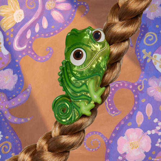 Tangled Pascal Hair Clip clasping a blonde braid, a shiny green chameleon-shaped barrette with large eyes from ColourPop Tools and Accessories, set against a whimsical purple floral painted backdrop.