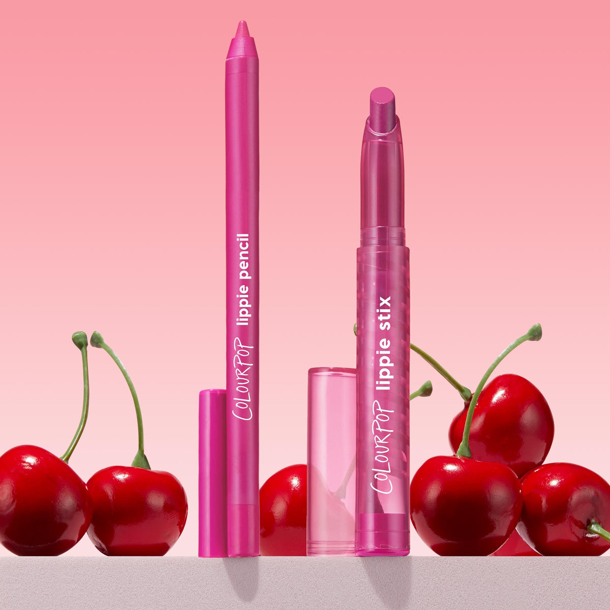 Heart On ColourPop Lippie Pencil stands upright with exposed tip in vibrant fuchsia, beside matching Lippie Stix and clear caps, surrounded by glossy red cherries against a pink gradient backdrop, showcasing precise, creamy, long‑wear lip liner.