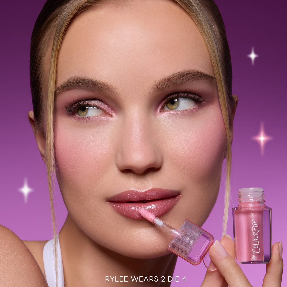 Model applies Gloss Me Up ultra glossy lip set in Heart to Get, holding the pink shimmery ColourPop-style tube against a purple starry background, showcasing high-shine nude-pink lips and matching soft glam makeup for a glossy finish.