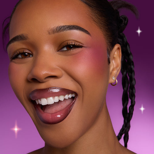 Smiling model wearing Heart to Get Sorry Not Sorry blush and UGL set, showing bold berry cheek color and glossy lips against a vibrant purple background with subtle sparkles.