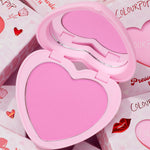 Heart-shaped Hot 2 Touch compact opens to reveal bright pink ColourPop Pressed Powder Blush, set in a pastel pink case with built-in mirror, resting atop Valentine-themed boxes with hearts and red lip graphics.