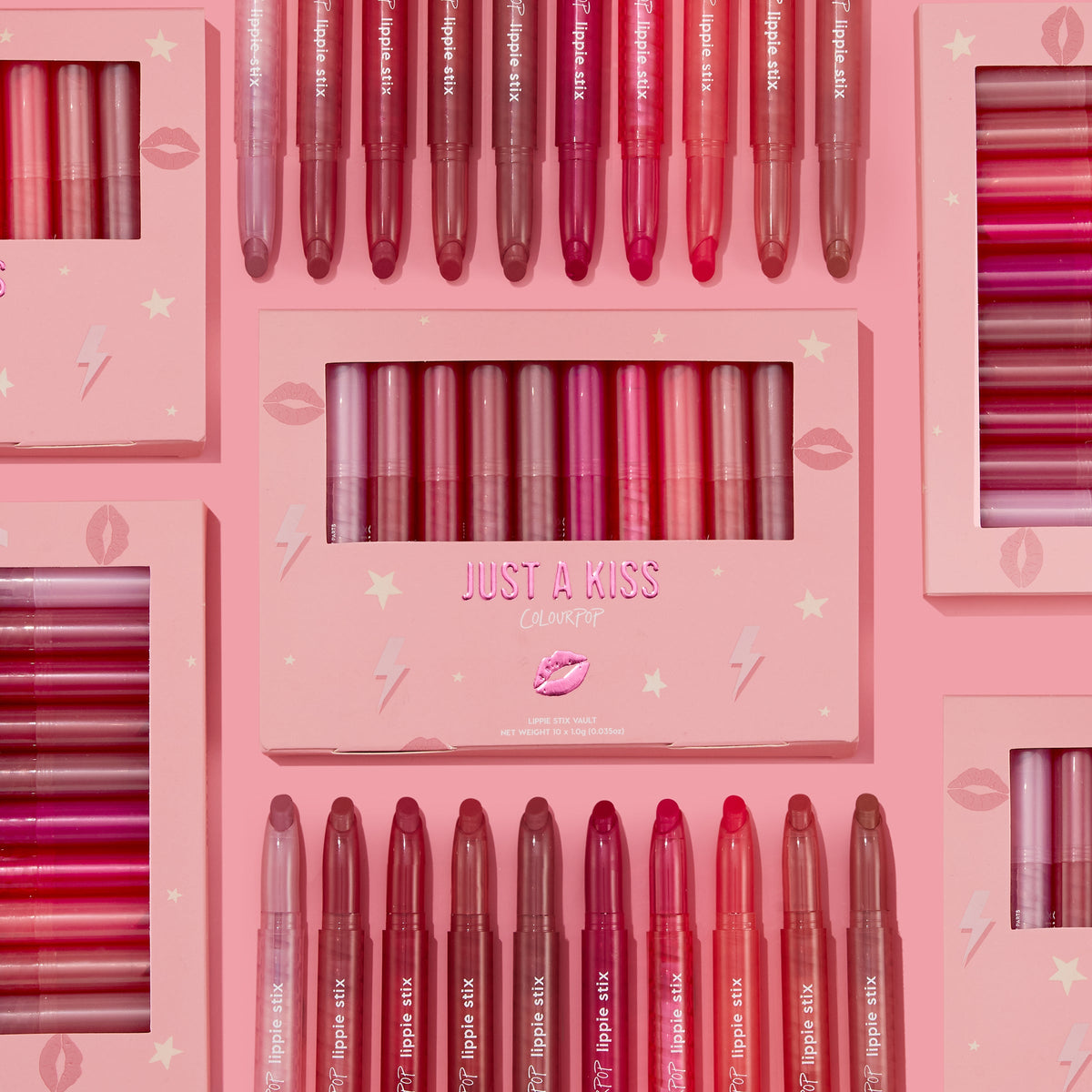 ColourPop Lippie Stix Just A Kiss set showcases lip crayon shades - pink, mauve, and red - arranged in rows with the boxed kit centered on a blush-pink background decorated with lips and lightning icons, gift-ready makeup vault.