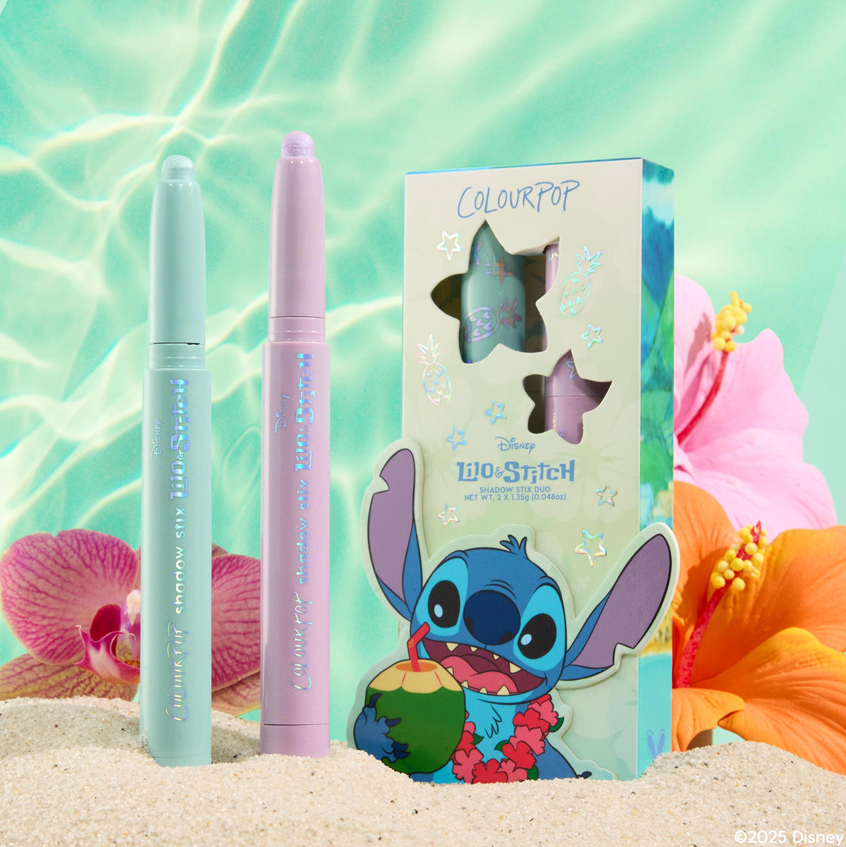 Mint and lavender ColourPop Shadow Stix Cute and Feisty duo stand uncapped with shimmery tips beside a Lilo & Stitch box with star cutouts, set in beach sand with pink orchids, orange hibiscus, and aqua water reflections.