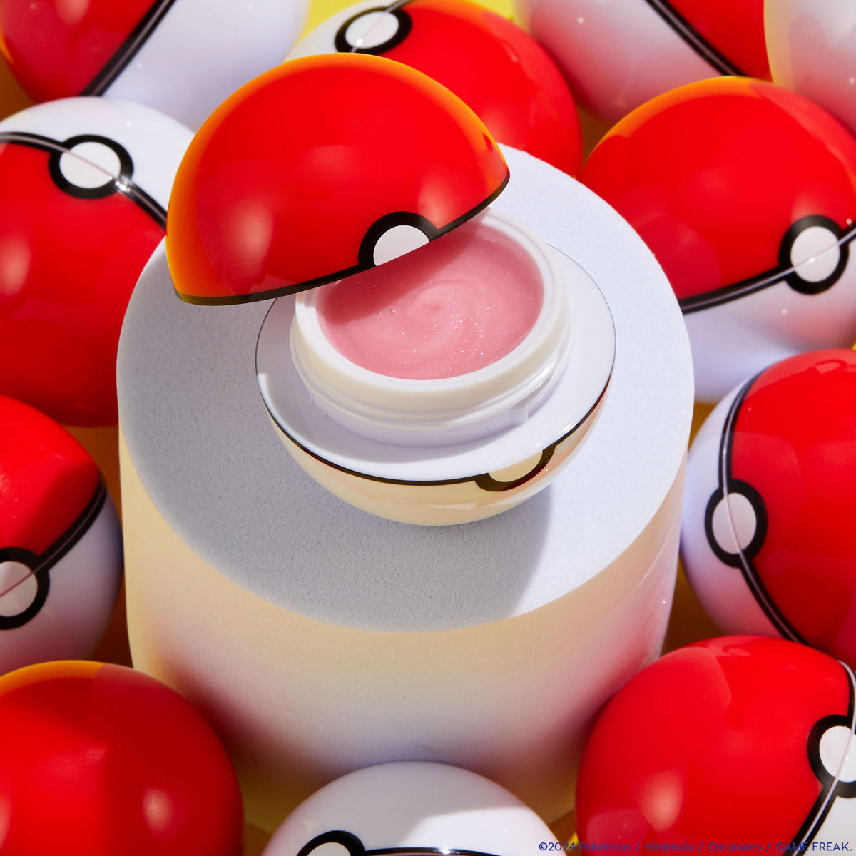 You're a Catch ColourPop Lip Mask - pink, glossy balm - sits open in a Poké Ball–style red-and-white container on a pastel pedestal, surrounded by matching spheres under bright studio light, highlighting the playful packaging and lip care product.